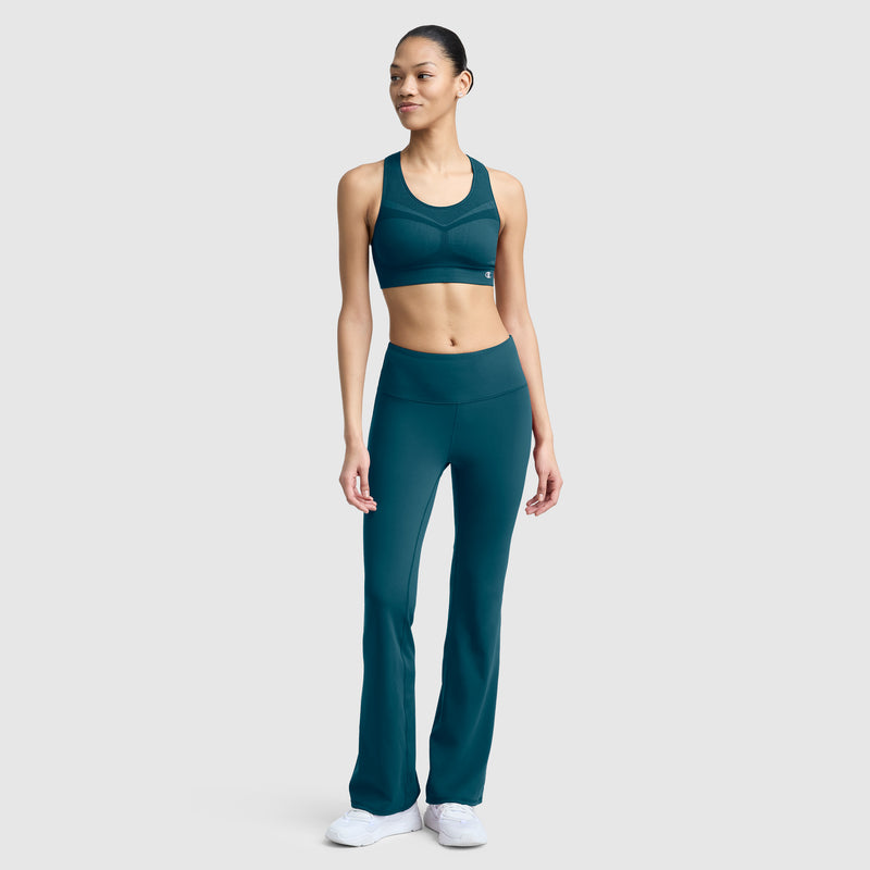 A woman wearing Champion’s Soft Touch Flare Leggings with Anti Odor technology and a C Logo stands and smiles slightly, looking to the side. She pairs the teal leggings with a matching sports bra and white sneakers. The plain light gray background highlights her stylish workout leggings for women.