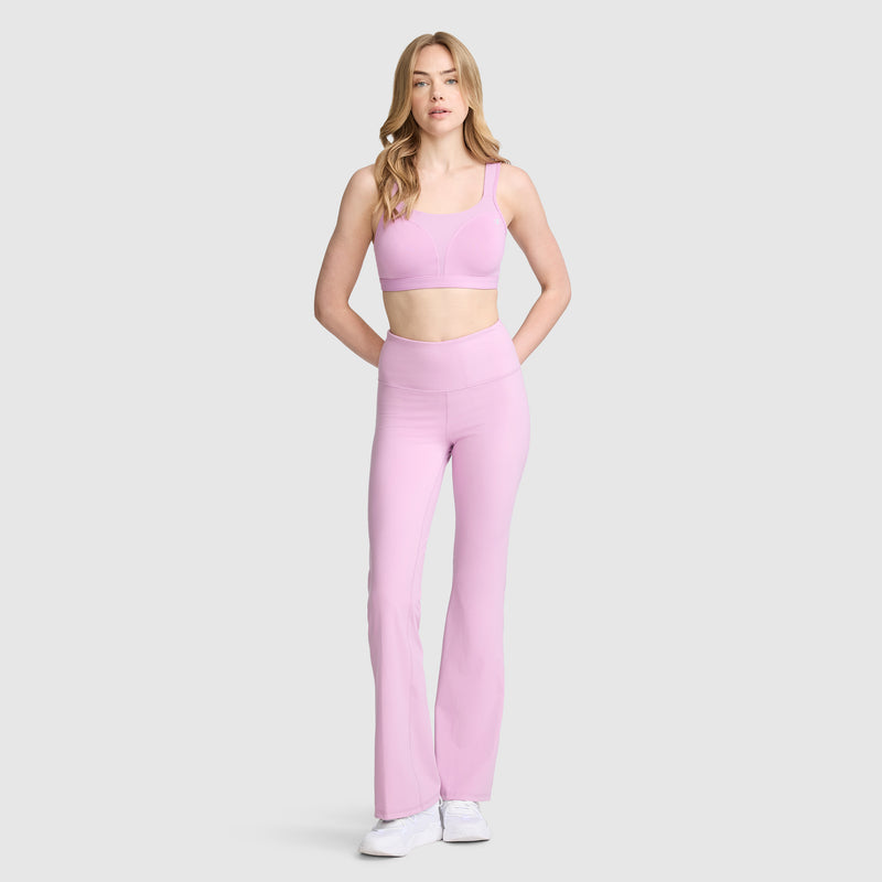 A woman stands against a plain background, wearing Champion’s Soft Touch Flare Leggings with the C Logo—anti-odor, high-waisted workout leggings in light pink—paired with a matching sports bra and white sneakers. Her hands are behind her back.