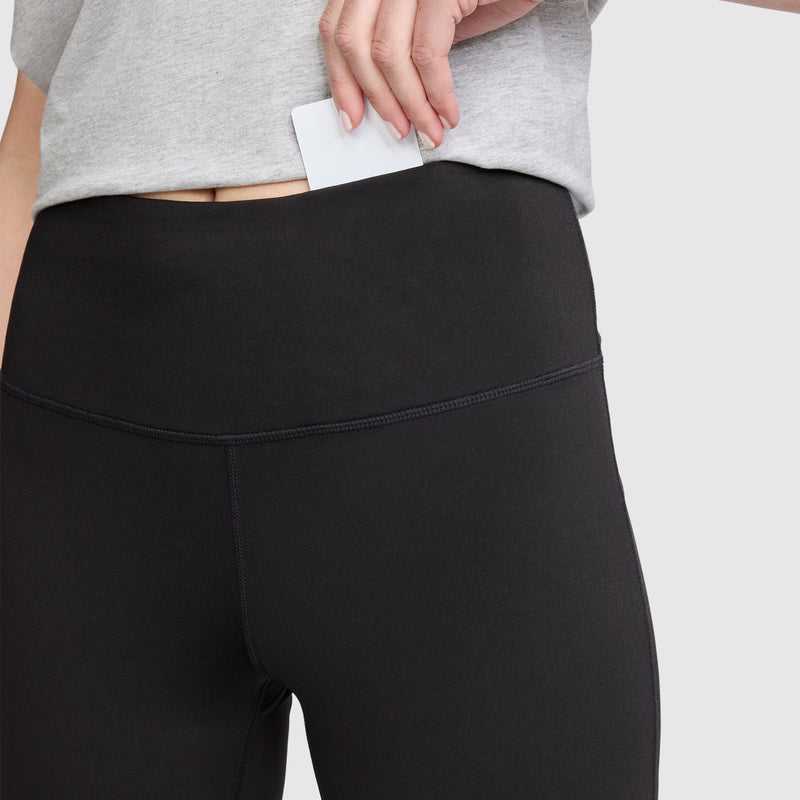 A person wearing Champion Soft Touch Flare Leggings with anti-odor technology and a C logo in black, paired with a gray shirt, places a white card into the hidden waistband pocket.