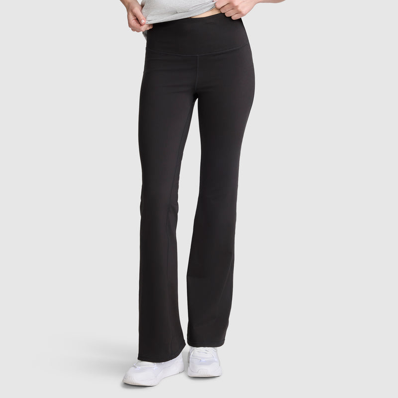 A person wearing Champion Soft Touch Flare Leggings with anti-odor technology and C logo in black, paired with white sneakers, stands against a plain light gray background. The upper body is partially visible, and their hands are adjusting the waistband of these workout leggings for women.