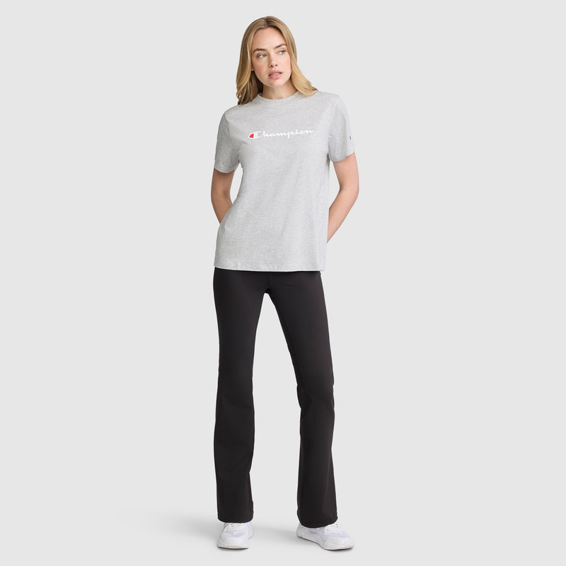 A woman stands facing forward against a plain light background, wearing a light gray Champion t-shirt and black Champion Soft Touch Flare Leggings with an anti-odor finish and C logo. She has her hands in her pockets, a neutral expression on her face, and completes the look with white sneakers.