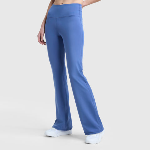 Someone wearing Champion's Soft Touch Flare Leggings—high-waisted, blue with anti-odor fabric, wide waistband, and signature C logo—paired with white sneakers stands against a plain light gray background. Torso not shown.
