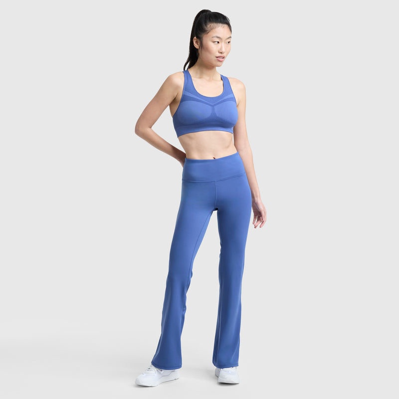 A woman stands against a light gray background wearing Champion Soft Touch Flare Leggings with a C Logo and anti-odor feature, paired with a matching blue sports bra and white sneakers. Her hair is in a ponytail and she has a neutral expression.