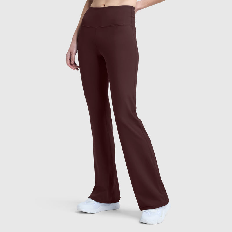 A person stands against a plain light gray background, wearing Champion Soft Touch Flare Leggings in a high-waisted burgundy style featuring anti-odor technology and the signature C logo, paired with white sneakers. Their upper body and face are not visible.