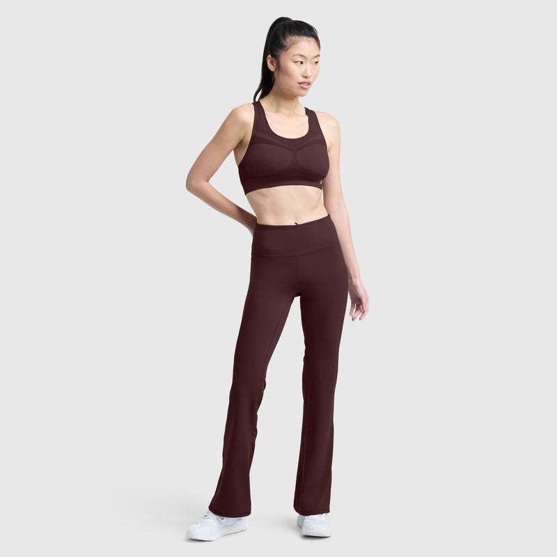 A woman stands against a plain background wearing Champion’s Soft Touch Flare Leggings with a C Logo in burgundy, paired with a matching sports bra and white sneakers. Her hair is pulled back in a ponytail as she looks to the side, highlighting these anti-odor, stylish women’s workout leggings.