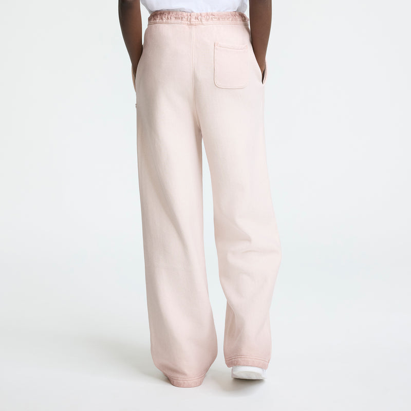 A person is seen from behind against a plain white background wearing Champion’s Time Capsule Vintage Loose Fit Pant in light pink with a relaxed fit, elastic waistband, wide legs, and a back pocket.