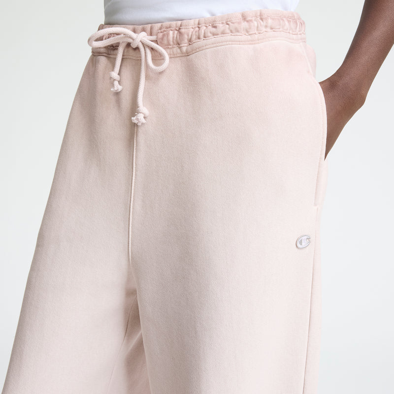 Close-up of a person wearing Champion Time Capsule Vintage Loose Fit Pant in light pink, featuring an elastic drawstring waistband, small embroidered logo on the side, relaxed fit, and one hand in the pocket. Made from soft Reverse Weave fleece.