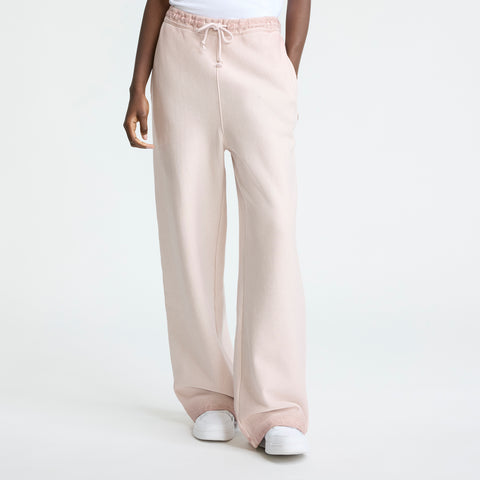 A person wears loose, light pink Champion Time Capsule Vintage Loose Fit Pants with a drawstring waist and white sneakers, standing against a plain white background. The image shows only the lower half, highlighting the relaxed fit.