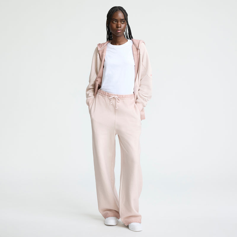 A woman stands against a white background wearing a white tee, light pink zip-up hoodie, and Champion Time Capsule Vintage Loose Fit Pant with white sneakers. Her hands in her pockets, she looks confidently ahead.
