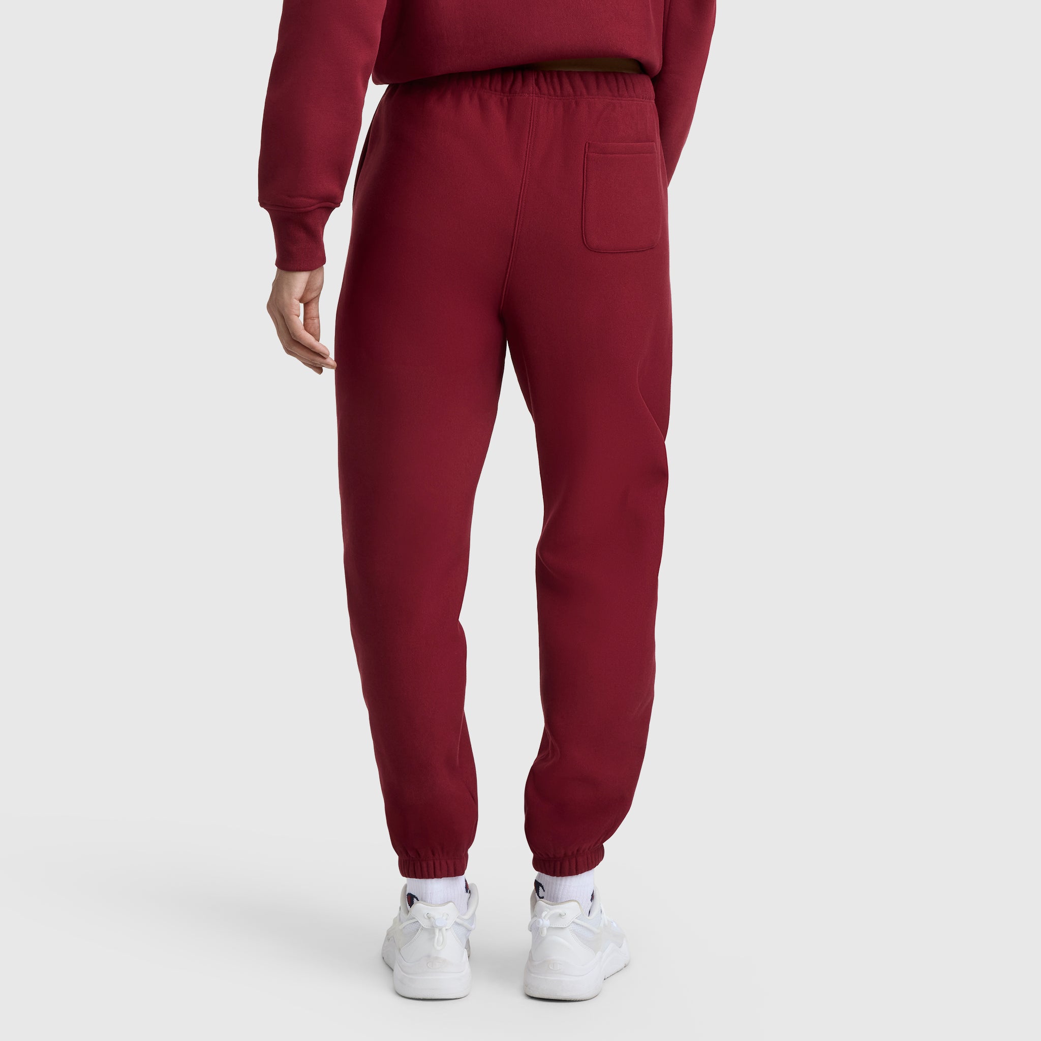 Someone is shown from the back wearing maroon Champion Essentials Reverse Weave Cinched Bottom Sweatpants and a matching sweatshirt, standing on a white surface with visible white sneakers.