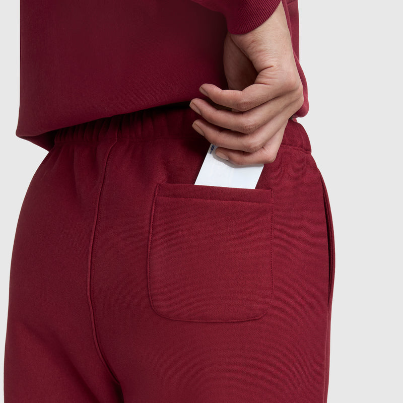 A person wearing Champion Essentials Reverse Weave Cinched Bottom Sweatpants in red holds a smartphone halfway inside the back pocket, their hand resting on the phone against a plain, light-colored background.