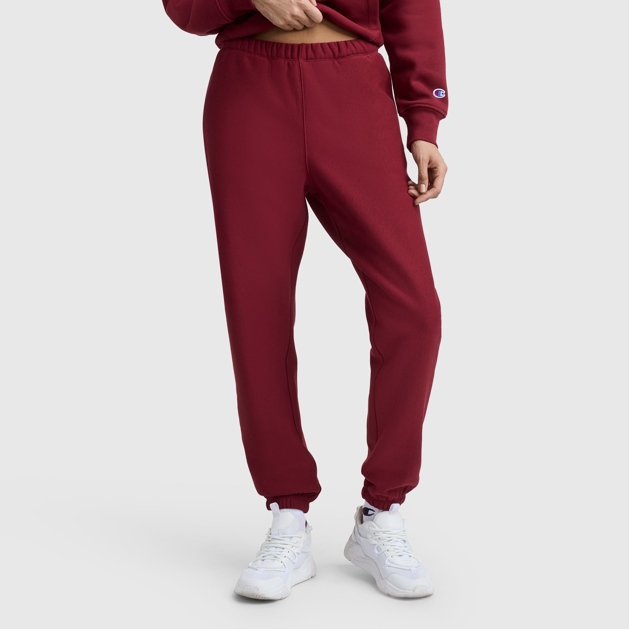 A person wearing maroon Champion Essentials Reverse Weave Cinched Bottom Sweatpants with a matching sweatshirt and white sneakers stands against a plain light gray background, showing only the lower half of the body.