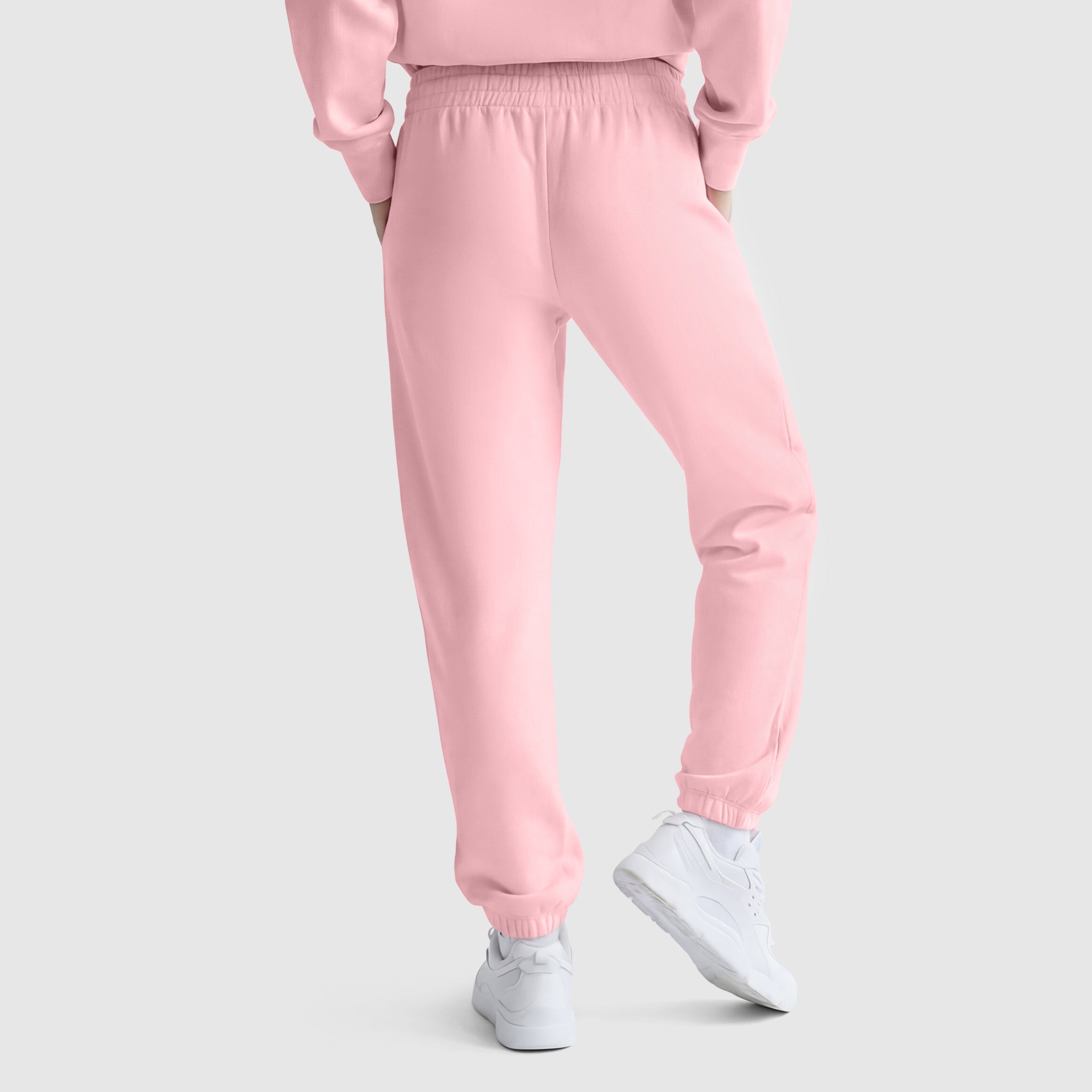 A person is shown from the back in light pink Champion Powerblend Oversized Sweatpants with C Logo, paired with a matching top and white sneakers. Hands in pockets, the look stands out against a plain light gray background.