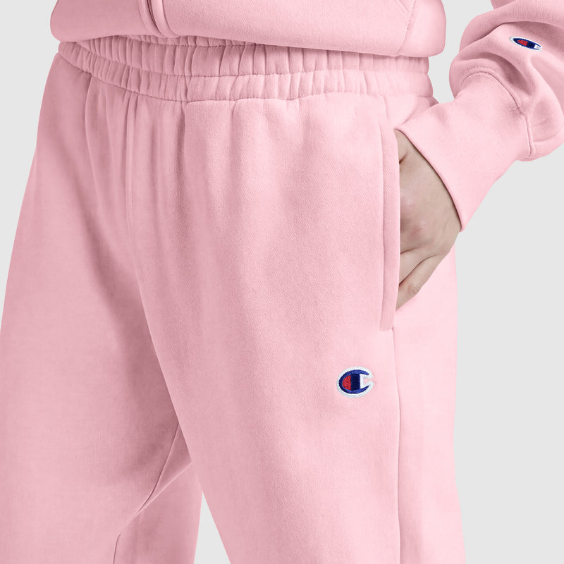 A person wears light pink Champion Powerblend Oversized Sweatpants with a visible C logo and a matching sweatshirt, standing with one hand in their pocket against a plain light gray background, showcasing this cozy women's loungewear look.