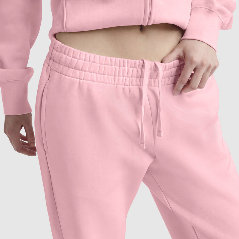 Model wears Champion Powerblend Oversized Sweatpants, C Logo in light pink fleece with a matching cropped hoodie, standing with one hand on hip and the other by their side, shown from shoulders to thighs against a light background.