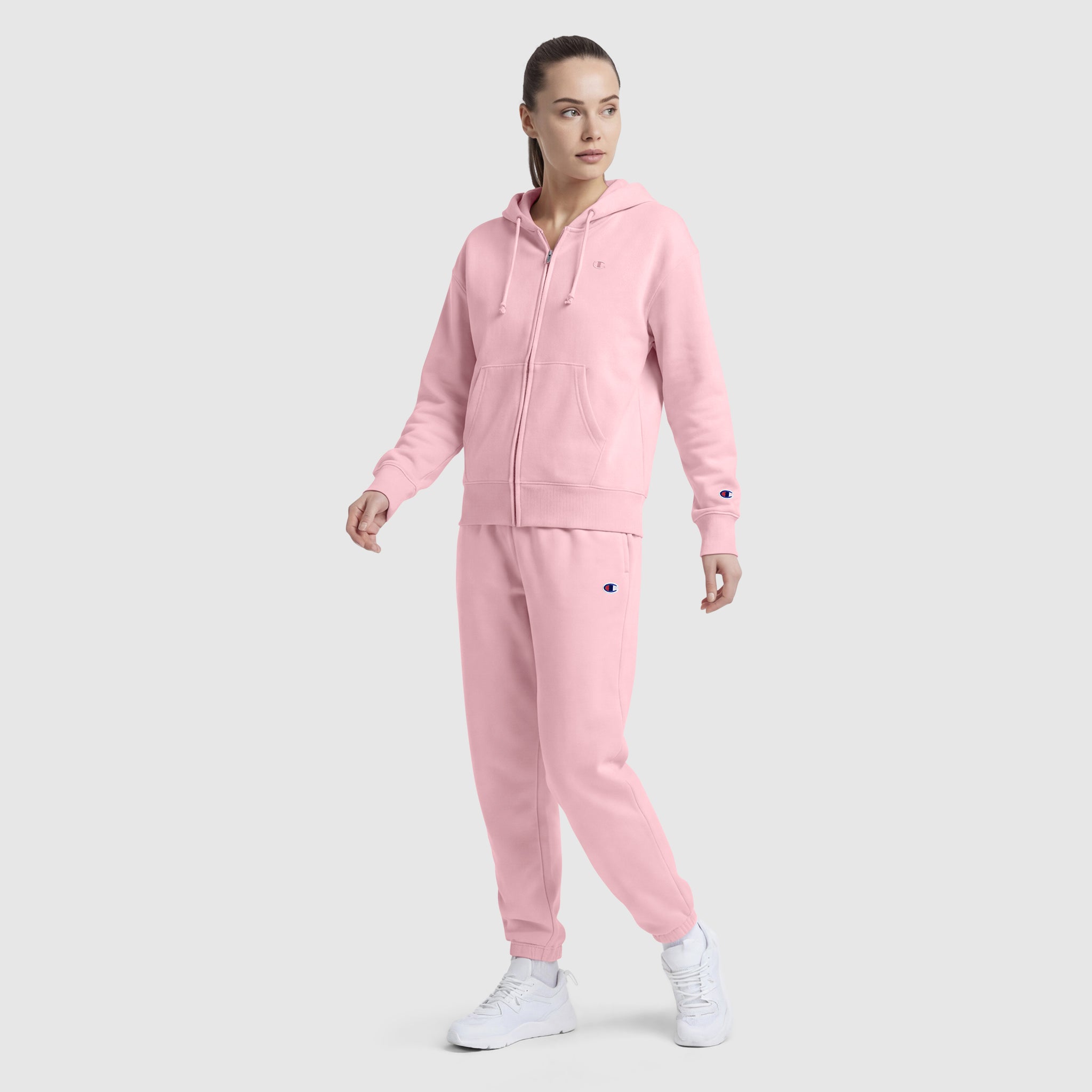 A woman wears Champion’s Powerblend Oversized Sweatpants with a C Logo in light pink, paired with a matching zip-up hoodie and white sneakers, posing against a plain light gray background.