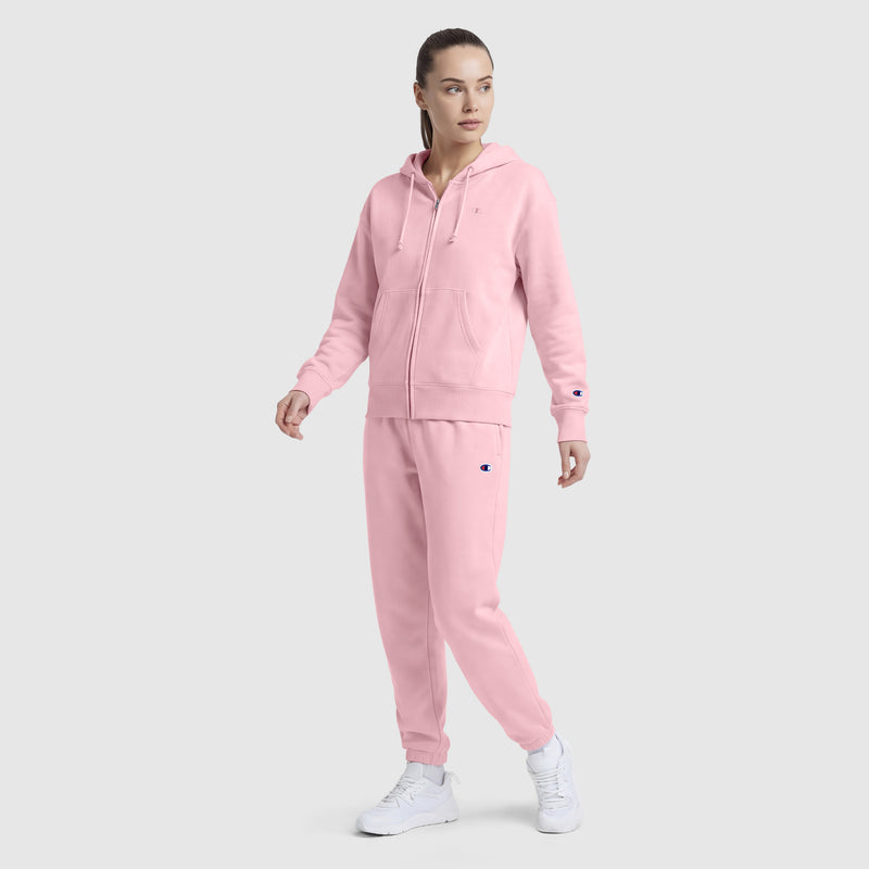 A woman wears Champion’s Powerblend Oversized Sweatpants with a C Logo in light pink, paired with a matching zip-up hoodie and white sneakers, posing against a plain light gray background.