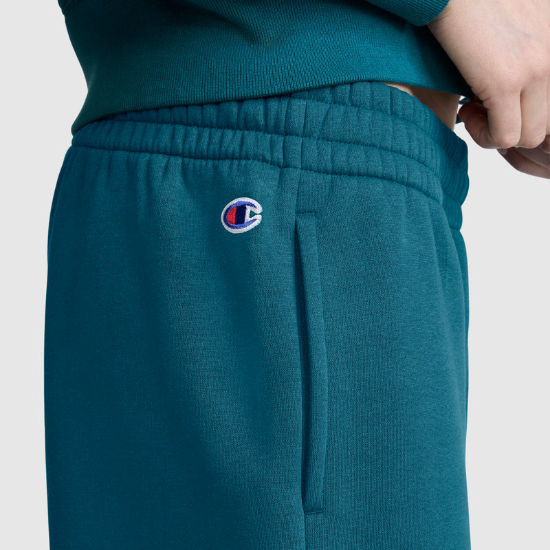 A close-up shows a person wearing teal Champion Powerblend Oversized Sweatpants, featuring an elastic waistband and a signature C logo above the pocket. Their shirt complements the Champion sweatpants, and their hand is resting near the waist.