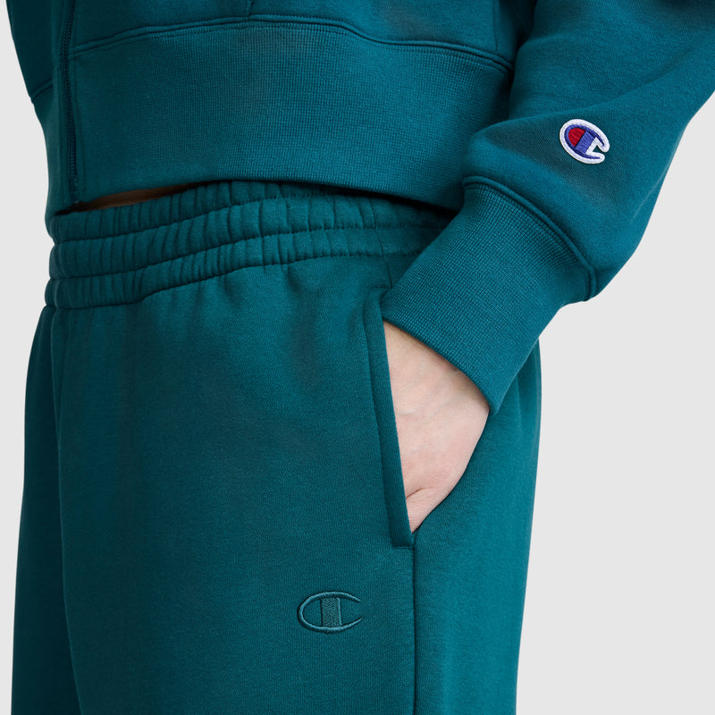 A person is wearing a teal Champion hoodie and Powerblend Oversized Sweatpants, C Logo by Champion, with their hand in the pocket. The Champion logo is visible on the sleeve and near the pocket.