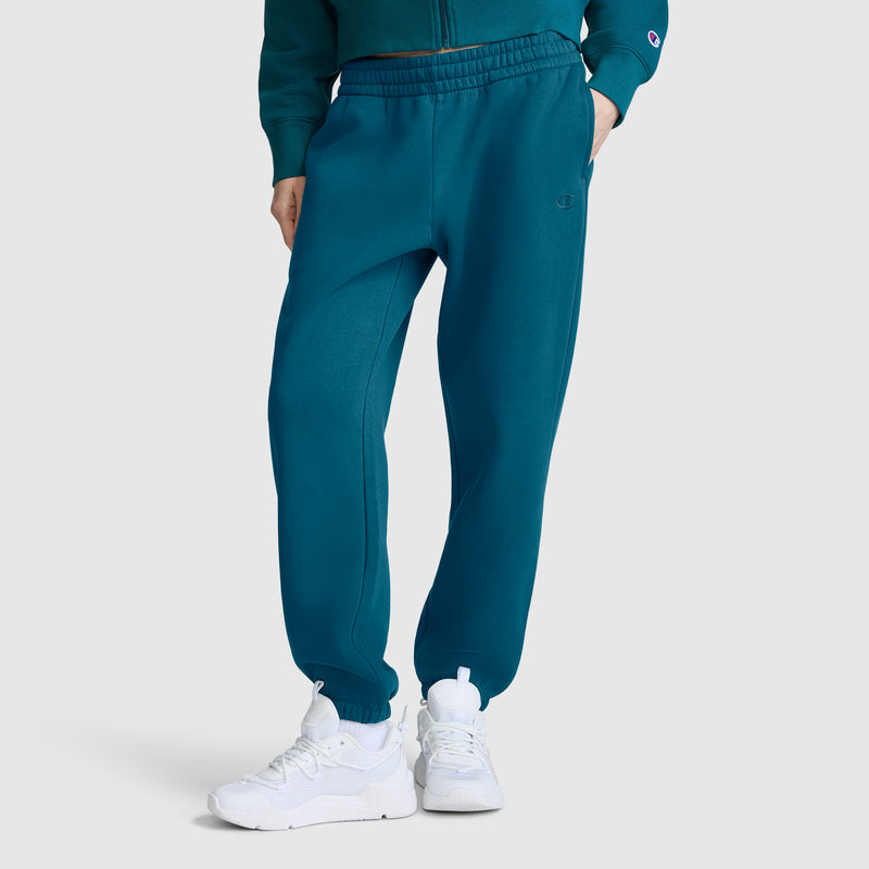 Model wearing Champion Powerblend Oversized Sweatpants, C Logo in teal, paired with white sneakers and a matching teal sweatshirt. Stands against a plain light background with upper body partially visible.