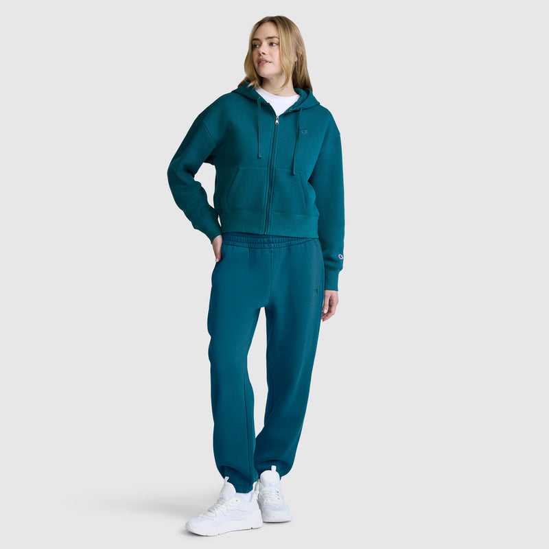 A woman stands against a plain light gray background, dressed in a teal zip-up hoodie and Champion Powerblend Oversized Sweatpants with the C Logo, styled with white sneakers and a white turtleneck.