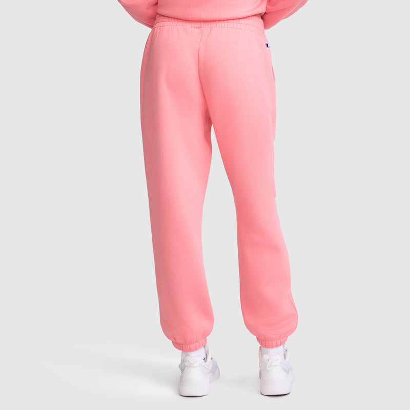 A person is shown from the back, standing against a plain light background, wearing Champion Powerblend Oversized Sweatpants with a C Logo in pink and white sneakers.