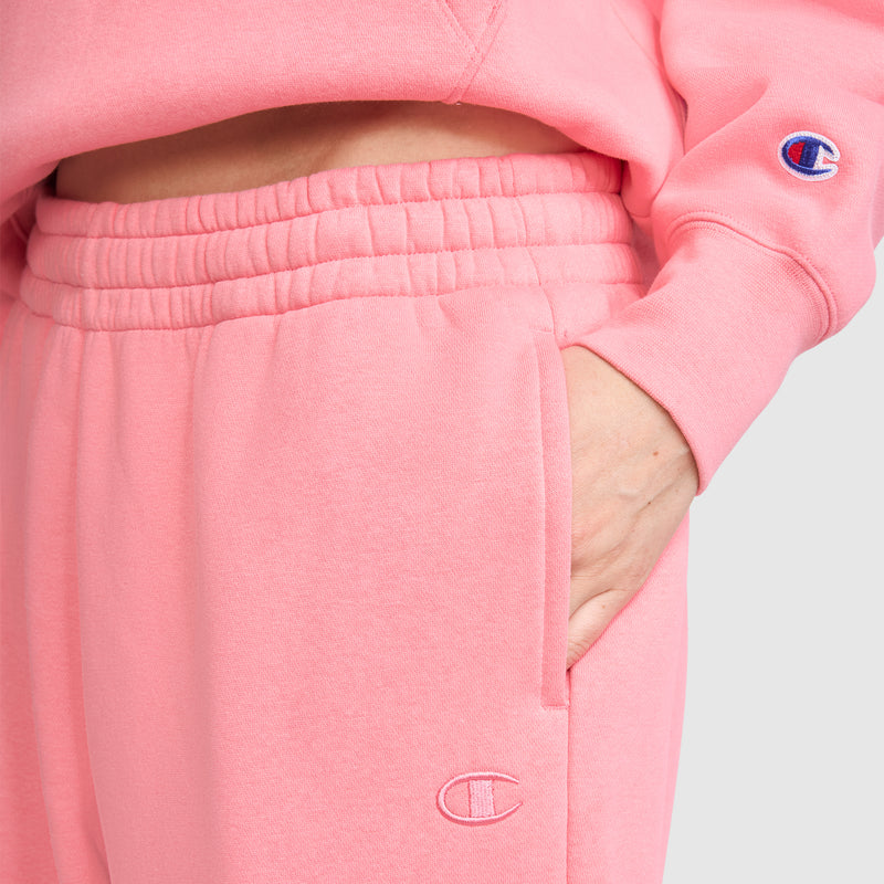 A person wearing a pink Champion Powerblend fleece sweatshirt and Champion Powerblend Oversized Sweatpants, C Logo, with one hand in the pocket. The Champion logo is visible on both the sleeve of the sweatshirt and on the pants.