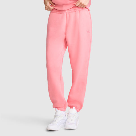 A person wearing light pink Champion Powerblend Oversized Sweatpants with the C Logo and white sneakers stands against a plain grey background. Only the lower half of the body is visible, with hands adjusting the waistband of the fleece pants.