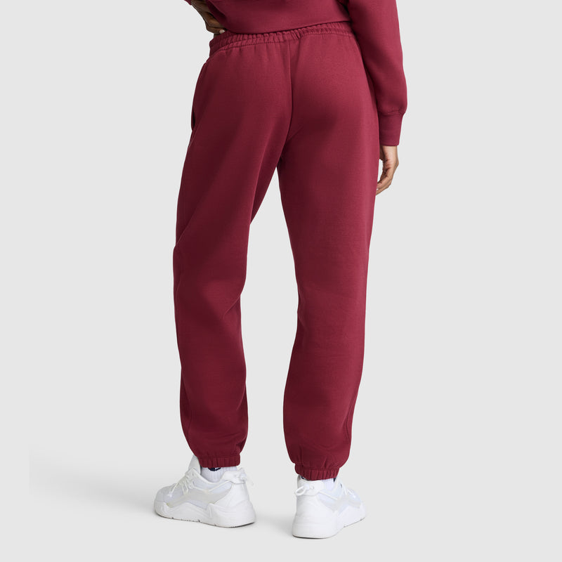 A person is shown from the back, wearing Champion Powerblend Oversized Sweatpants with the C Logo in maroon and white sneakers, standing against a plain light gray background. Only the lower half of the body is visible.