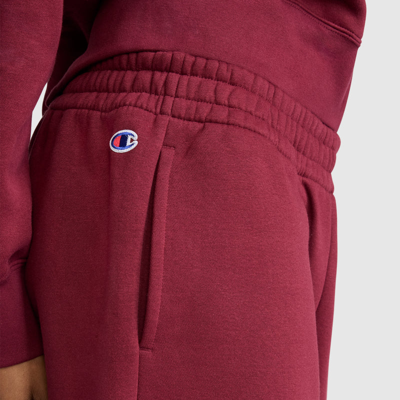 A woman is wearing Champion Powerblend Oversized Sweatpants with the C Logo in maroon, made from cozy fleece and designed with an elastic waistband and signature logo on the left pocket, paired with a matching maroon sweatshirt. The background is plain light gray.