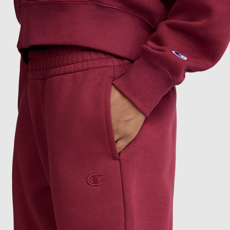 A person is wearing a matching maroon sweatshirt and Champion Powerblend Oversized Sweatpants with the signature C Logo, made from soft fleece. Their right hand rests in the sweatpants pocket, and the Champion logo is visible on both the sleeve and pants. The background is plain and light-colored.