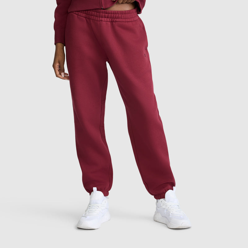 A person wearing maroon Champion Powerblend Oversized Sweatpants with the signature C Logo and a matching top stands with one hand by their side. The cozy fleece joggers combine comfort and style, paired with white sneakers. The background is a plain light gray, and the person’s head is not visible in the image.