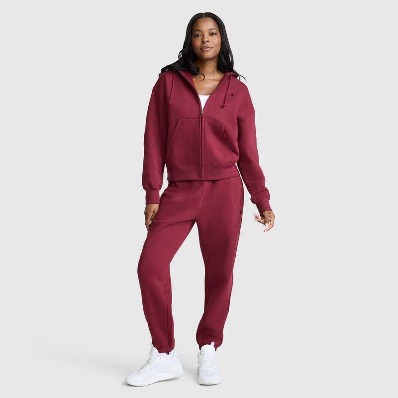 A woman stands wearing an oversized maroon zip-up hoodie and Champion Powerblend Oversized Sweatpants with the C Logo, paired with white sneakers, against a plain light gray background.