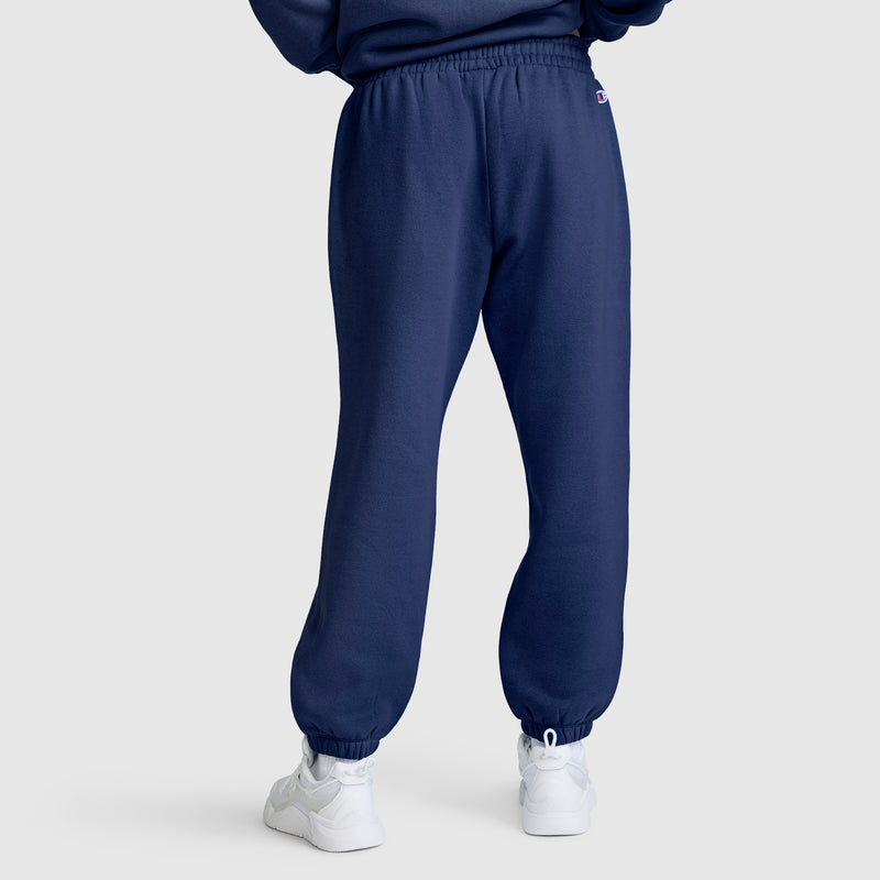 A person is wearing Champion’s Powerblend Oversized Sweatpants, C Logo in navy blue and white sneakers, standing on a light gray background. The image shows the back view of the sweatpants, which feature an elastic waistband and ankle cuffs.