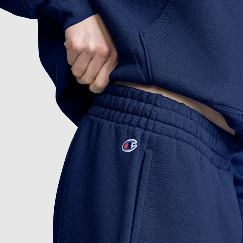 A person wearing navy blue Champion Powerblend Oversized Sweatpants, C Logo, and a matching hoodie adjusts the waistband of the pants. The Champion logo is visible on the left side of these cozy sweatpants.