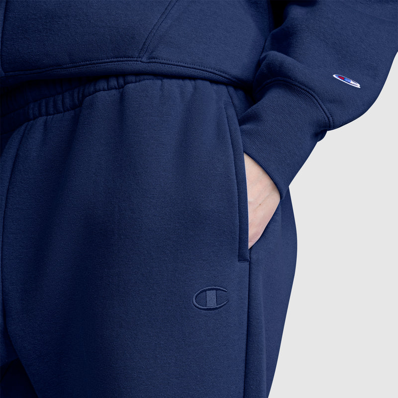 Close-up of a person wearing navy blue Champion Powerblend Oversized Sweatpants, C Logo, and a matching sweatshirt, with one hand in the pocket. The sweatpants feature the iconic Champion logo on both the pants and the sleeve of the sweatshirt.