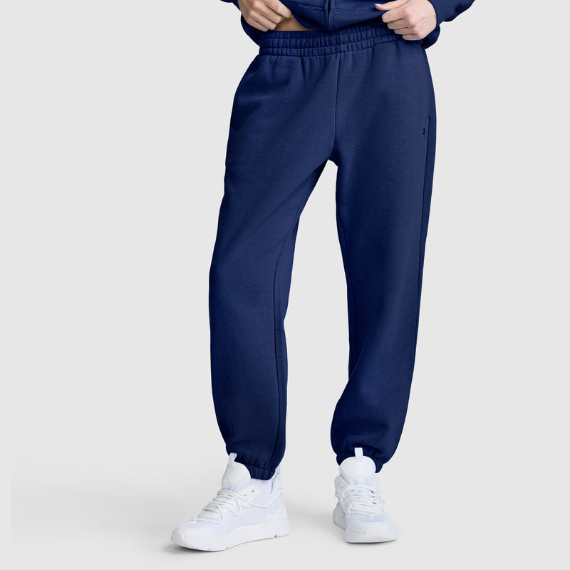 A person wearing Champion Powerblend Oversized Sweatpants with C Logo in navy blue and white sneakers, standing against a plain light gray background. Only the lower half of the body is visible.