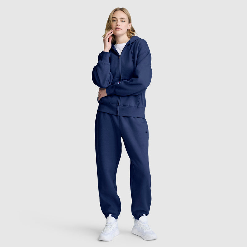 A woman stands wearing a navy blue zip-up hoodie, Champion Powerblend Oversized Sweatpants with the C Logo, and white sneakers against a plain light gray background. She poses with one hand touching her face and looks straight ahead.