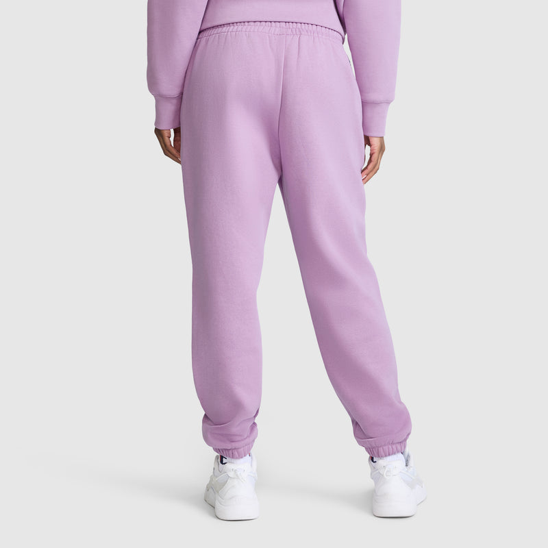 A person is seen from behind against a plain background, wearing Champion’s Powerblend Oversized Sweatpants with the C Logo in light purple, paired with a matching sweatshirt and white sneakers.