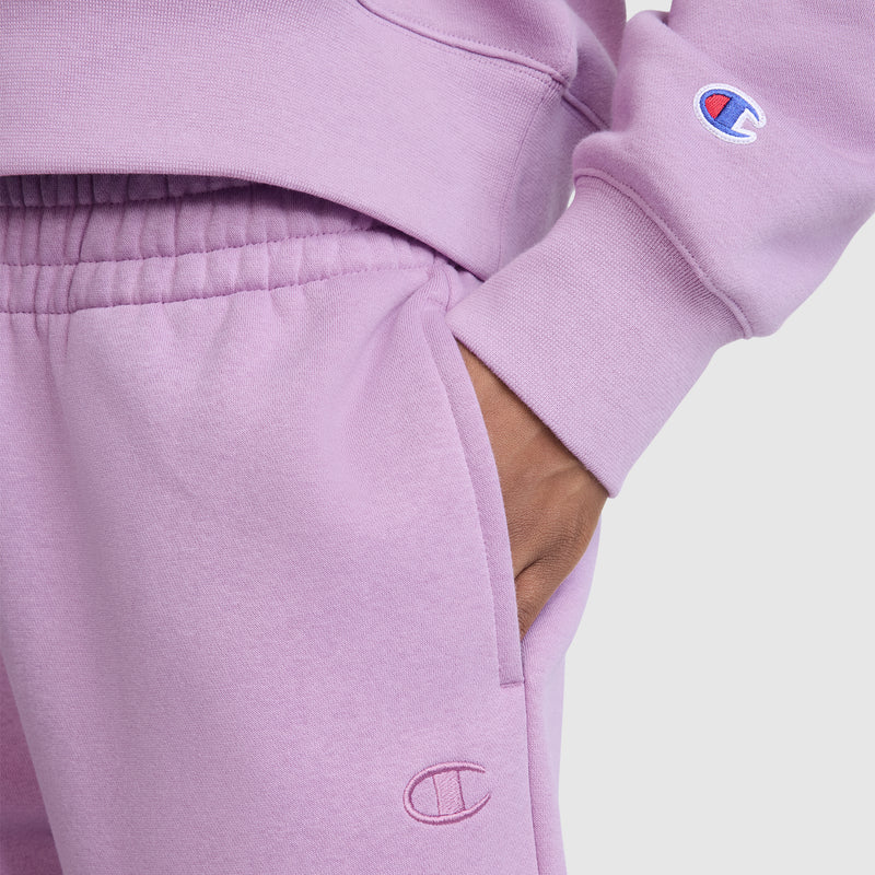 Wearing a matching lavender Champion sweatshirt and Champion Powerblend Oversized Sweatpants with the C Logo, the person stands with one hand in their pocket. Both garments showcase the iconic Champion branding and are made from soft Powerblend fleece for added comfort.