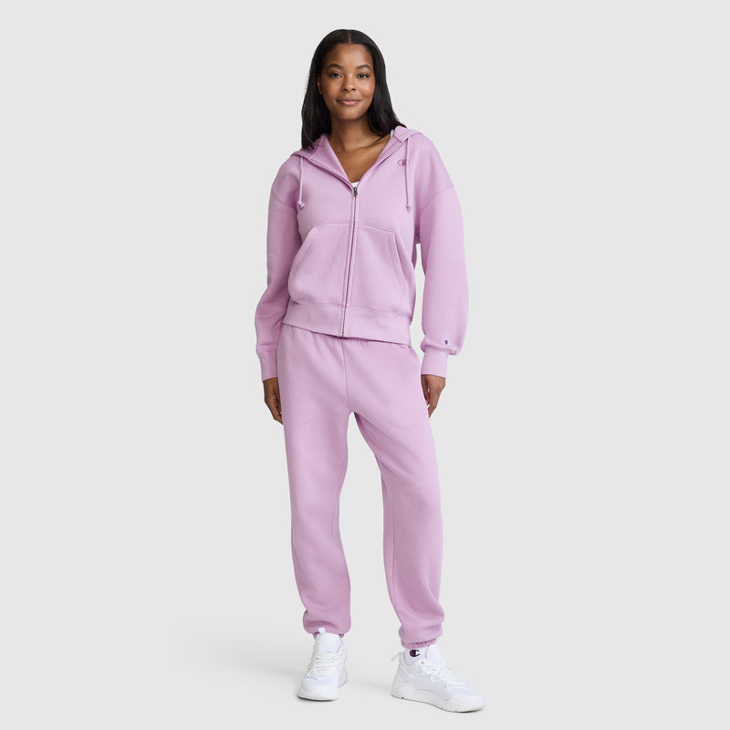 A person stands smiling against a plain light background, wearing white sneakers and Champion’s Powerblend Oversized Sweatpants with the C Logo, paired with a matching light purple zip-up hoodie.