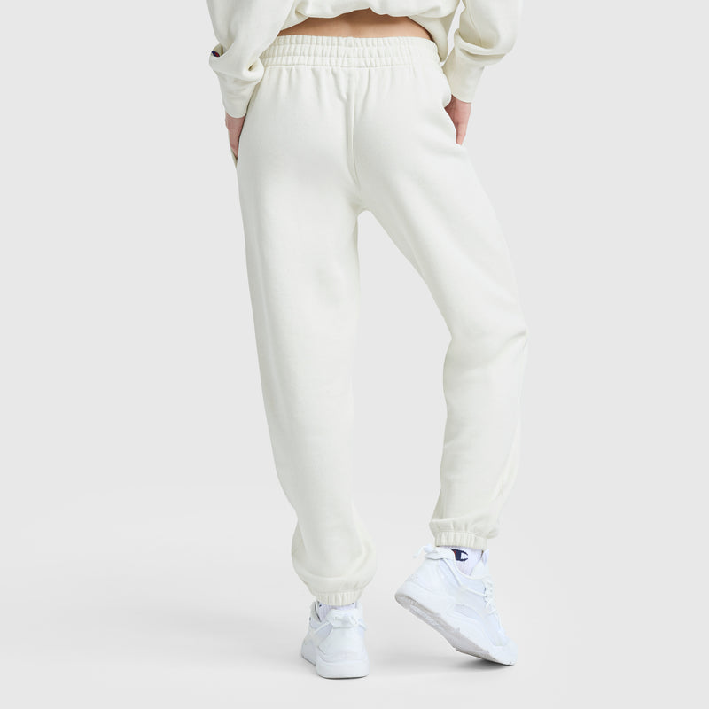 A person stands facing away, hands in pockets, wearing Champion Powerblend Oversized Sweatpants with a C Logo and a matching sweatshirt. They also have on white athletic sneakers. The background is plain and light-colored.