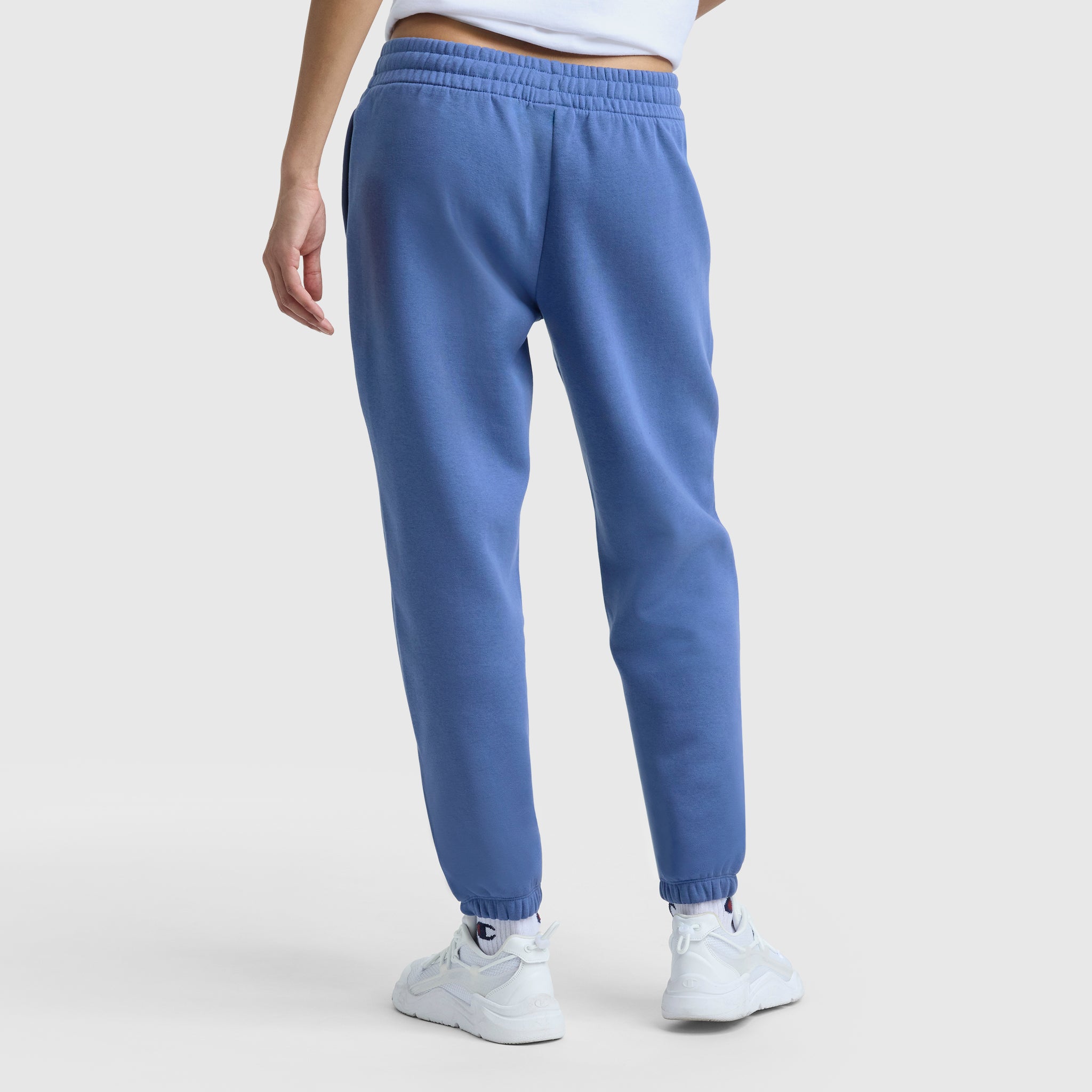 A person stands facing away, showing only the lower half of their body, wearing Champion Powerblend Oversized Sweatpants with the C Logo in blue and white sneakers against a plain light background.