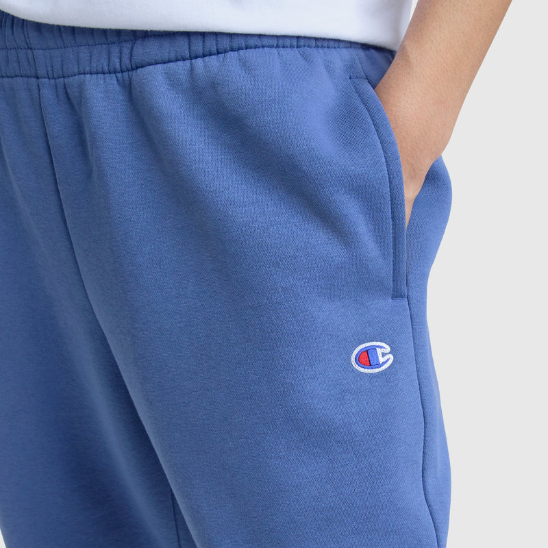 A person wears Champion Powerblend Oversized Sweatpants, C Logo, in blue with a hand in their pocket. The sweatpants have the embroidered Champion logo on the left thigh. The background is plain and light-colored.