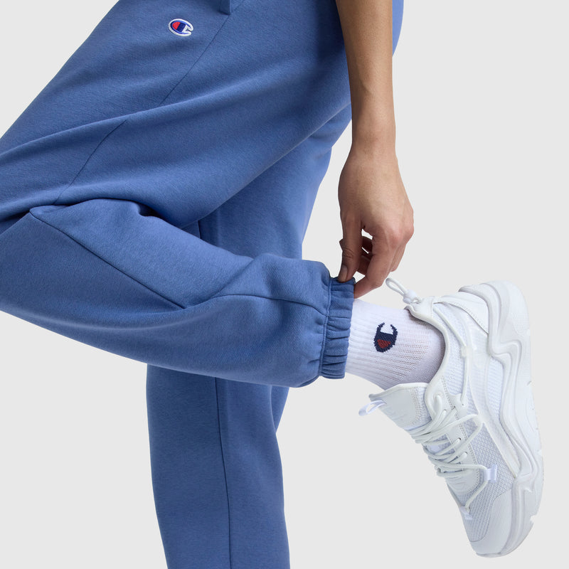 A person stands on one leg, holding their ankle, wearing Champion Powerblend Oversized Sweatpants with a C Logo and white socks with the Champion logo—perfect women's loungewear.