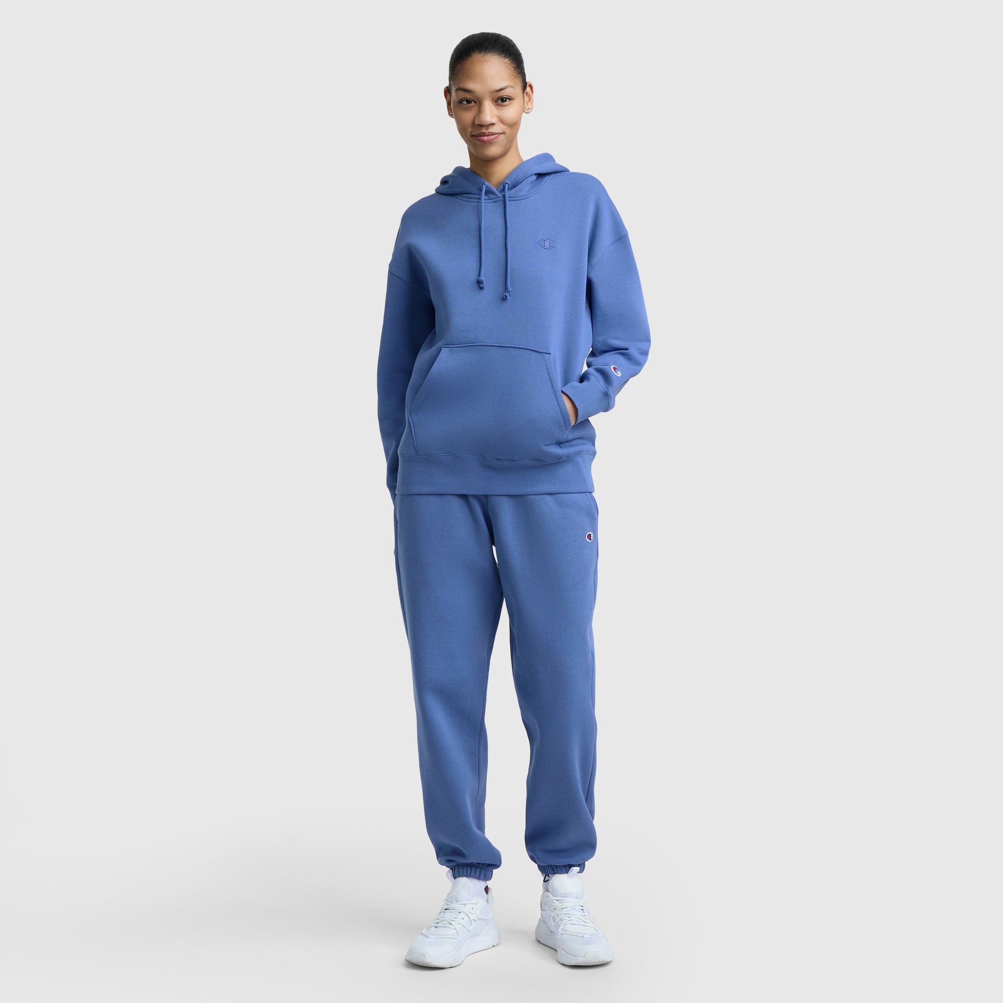 A woman faces forward against a plain white background, wearing Champion’s Powerblend Oversized Sweatpants with C Logo, paired with a matching blue hoodie and white sneakers—a perfect women’s loungewear look.