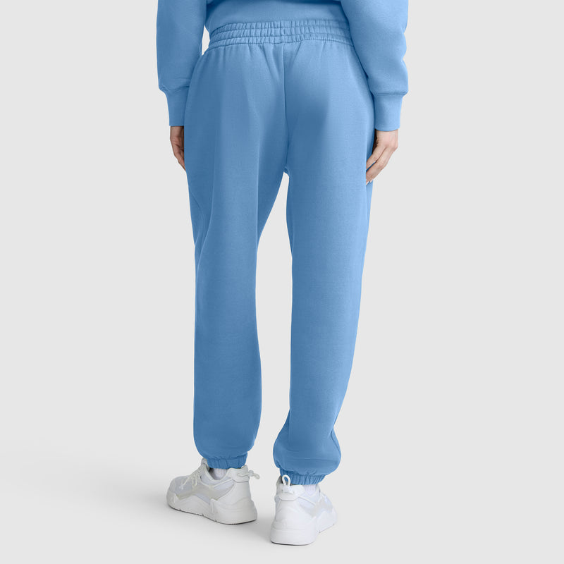 A person is shown from the back, standing against a plain white background, wearing Champion Powerblend Oversized Sweatpants with a C Logo in light blue, paired with a matching sweatshirt and white sneakers.