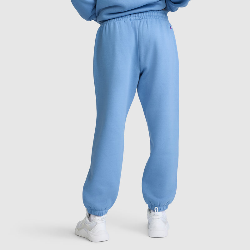 A person seen from the back stands on a plain white background, wearing light blue Champion Powerblend Oversized Sweatpants with the C Logo and white sneakers.