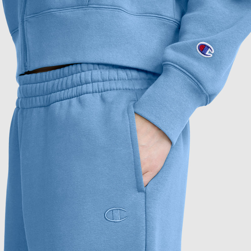 A person is wearing a light blue Champion Powerblend Oversized Sweatpants with the C Logo, their right hand tucked in the pocket. The Champion branding is visible on both the sleeve and pants, and the oversized sweatpants are made from soft Powerblend fleece for added comfort.