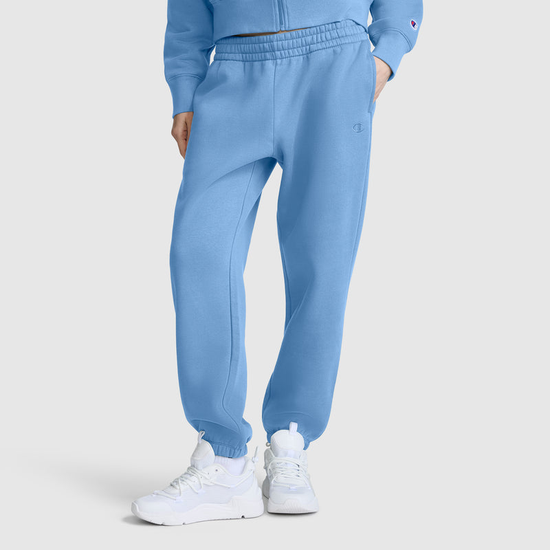 A person wearing Champion Powerblend Oversized Sweatpants with a C Logo in light blue, paired with a matching jacket and white sneakers, stands against a plain white background; only the lower half of the body is visible.